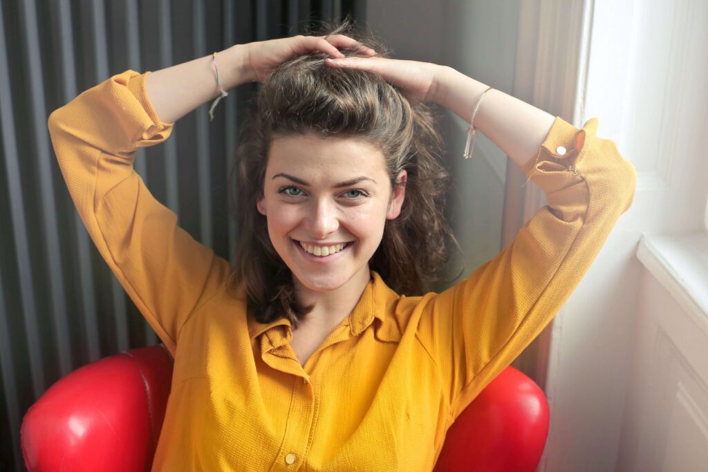 Smiling woman in yellow blouse sitting in red chair with hands in hair showing confidence and positive emotions. Find clarity about your strengths and challenges with psychological evaluations in Fort Collins, CO.