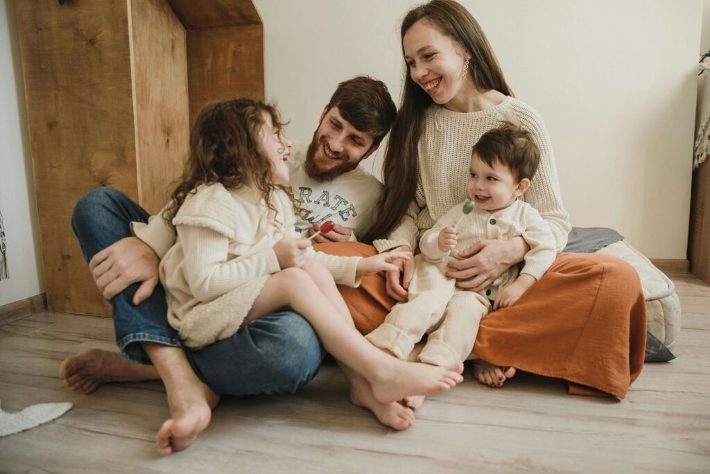 Happy family of four sitting together on bedroom floor laughing and playing, parents with two young children in cream outfits. Get the in-depth answers and individualized support plan your family needs with autism evaluations in Fort Collins, CO.
