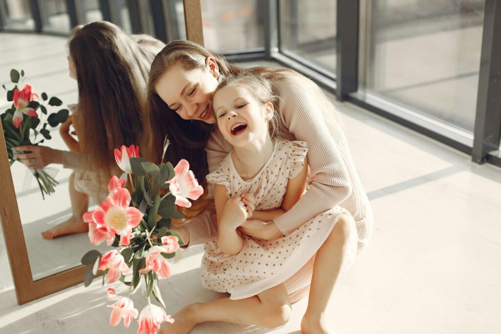 Mother and young daughter laughing together while looking in mirror with pink flowers in sunny room by window. Prepare with confidence and reduce stress for your child's appointment with autism evaluations in Fort Collins, CO.