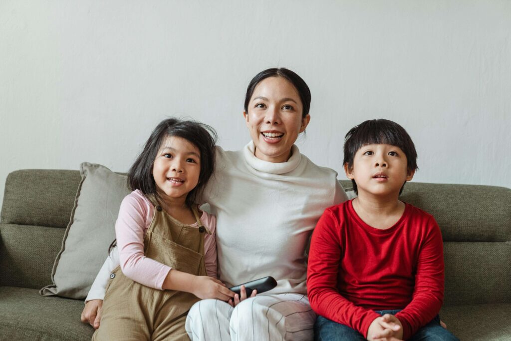 Smiling mother sitting on gray couch with two young children, daughter in beige overalls and son in red shirt, in bright living room. Find the support and guidance your family needs with autism testing for children in Fort Collins, CO.