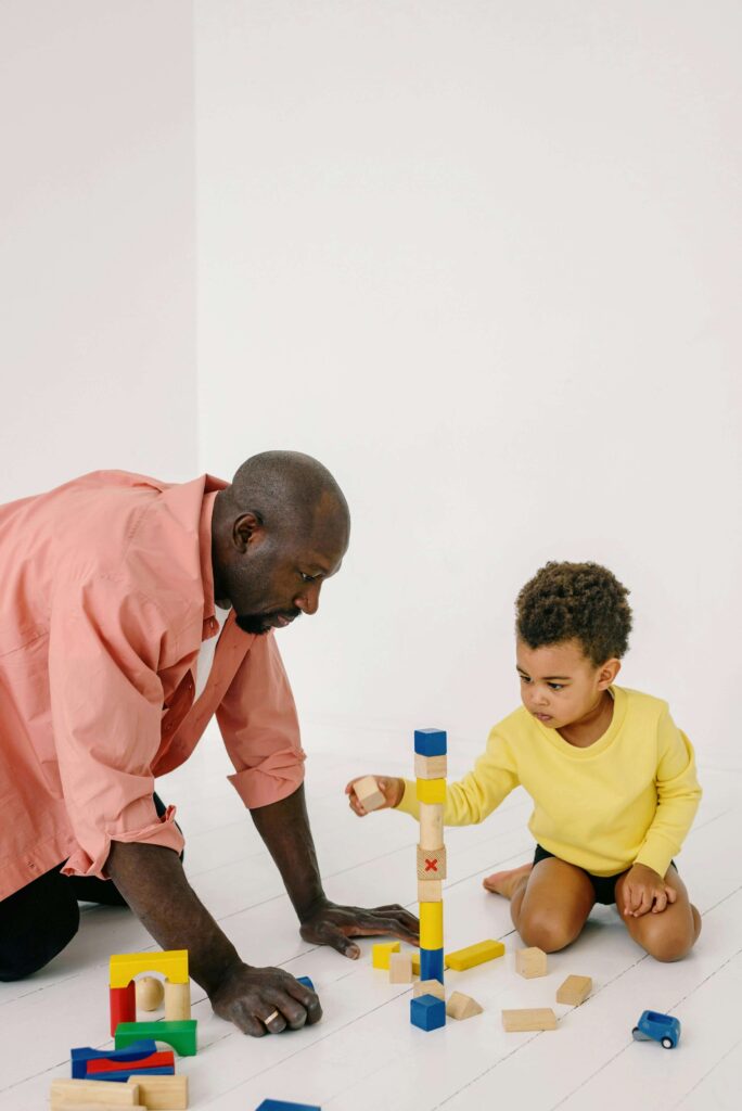 Father in pink shirt and young child in yellow shirt sitting on white floor building tower together with colorful wooden blocks. Learn the difference between screening and full assessment to get the right support with autism evaluations in Fort Collins, CO.