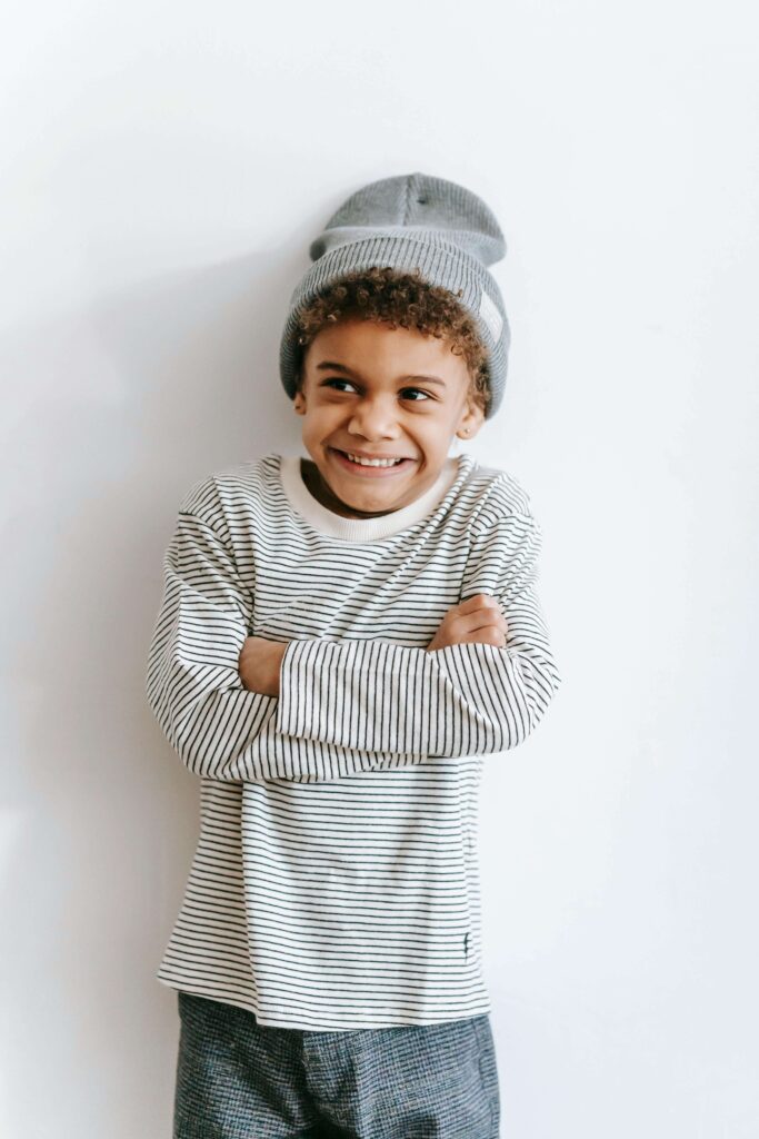 Smiling young boy in striped shirt and gray beanie with arms crossed against white background. Help your child feel comfortable and confident throughout the process with autism evaluations in Fort Collins, CO.