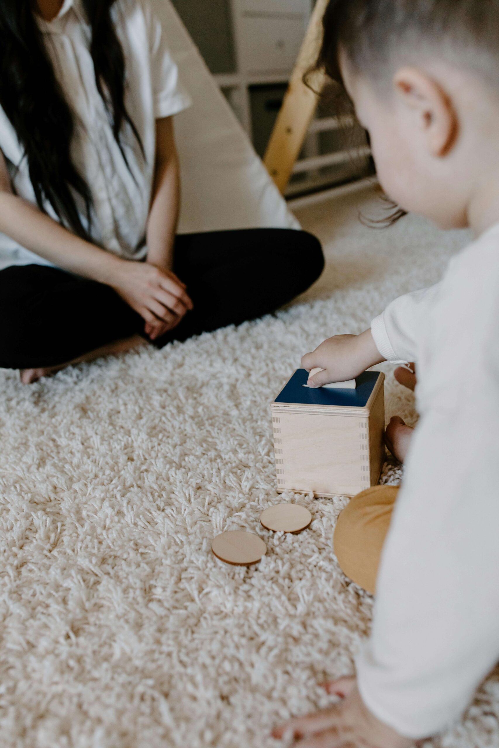 Mother sitting cross-legged on cream carpet watching toddler play with wooden shape sorter toy. Move beyond brief screenings to get diagnostic clarity and detailed recommendations with autism evaluations in Fort Collins, CO.
