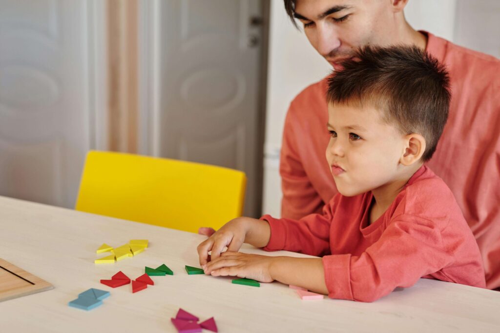 Father and young son in matching coral shirts working together on colorful geometric shape puzzle at table. Understand whether your child needs a screening or comprehensive assessment with autism evaluations in Fort Collins, CO.