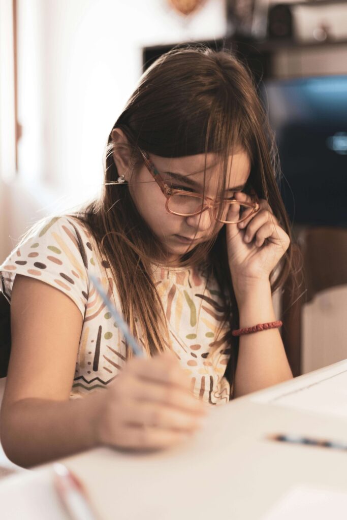 Young girl with glasses looking down at homework with stressed expression while writing at desk in bright room. Understand why your daughter struggles quietly despite working twice as hard with ADHD testing for kids in Fort Collins, CO.