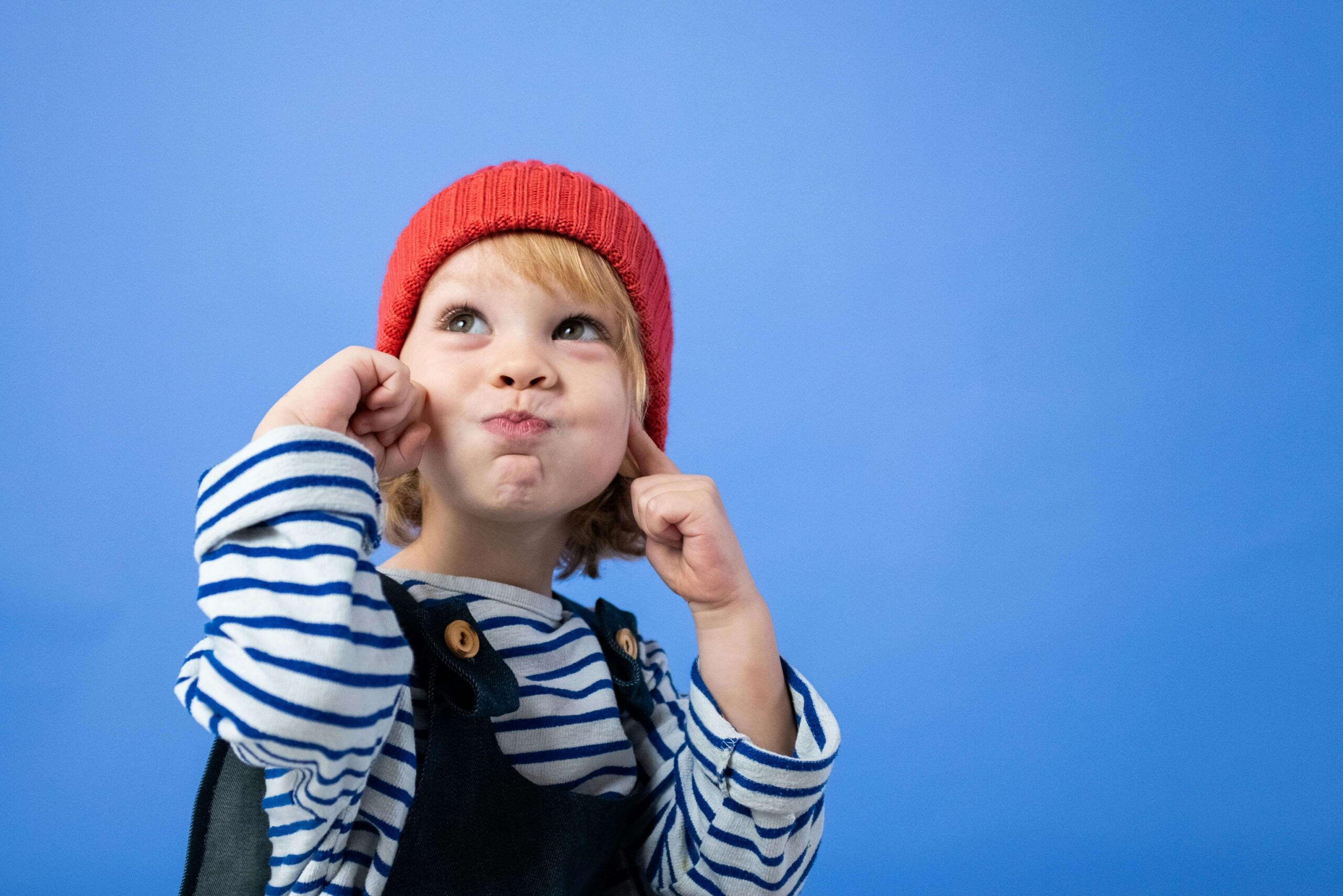 Young child in red beanie and striped shirt with hands on face making thoughtful expression against blue background. Discover how executive functioning impacts your child's daily life with comprehensive ADHD testing for kids in Fort Collins, CO.