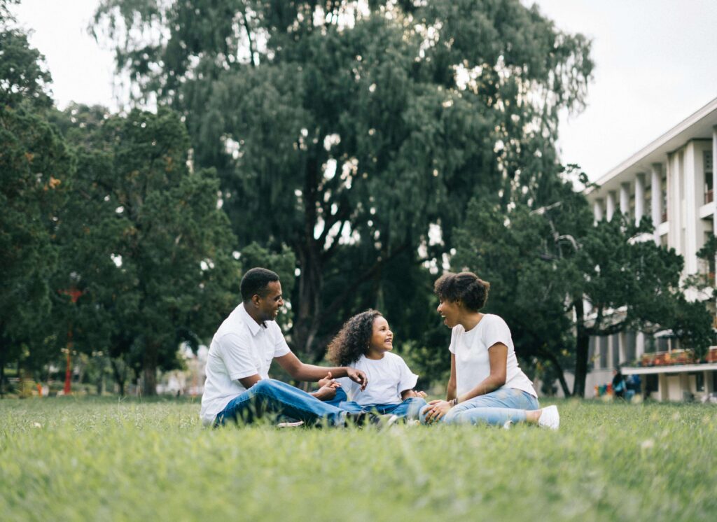 Happy family of three sitting together on grass in park with trees and building in background having conversation. Get the clarity your family needs about quiet ADHD symptoms with compassionate ADHD testing for kids in Fort Collins, CO.