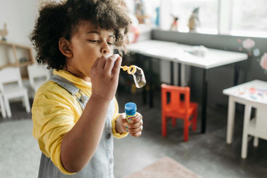 Young child in yellow shirt and overalls blowing bubbles in bright playroom with toys and furniture in background. Understand why your child struggles with organization and task completion through ADHD testing for kids in Fort Collins, CO.